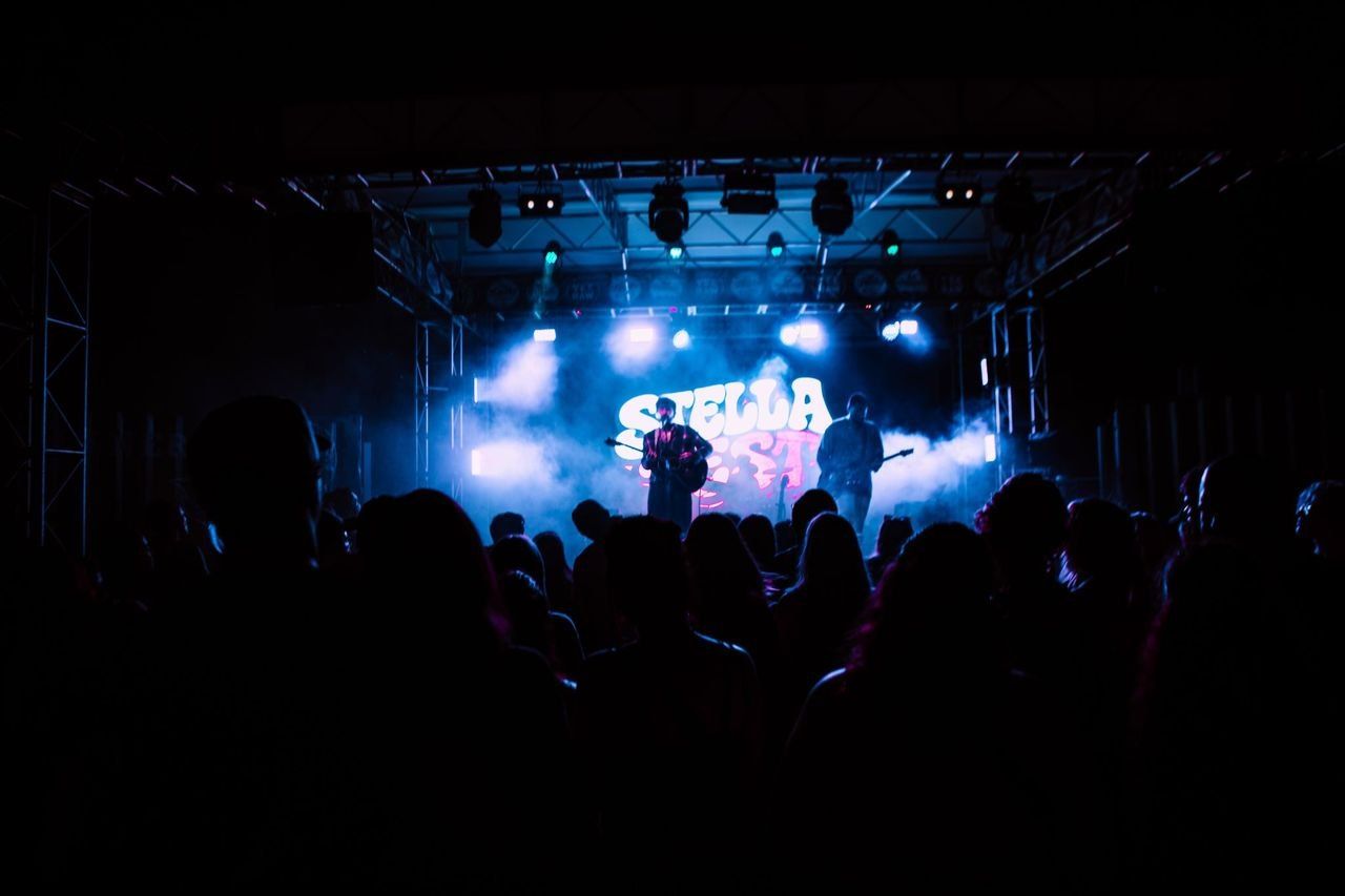 The Stella Fest stage lit in blue, two performers playing beneath the festival backdrop, seen from the back of a full room.